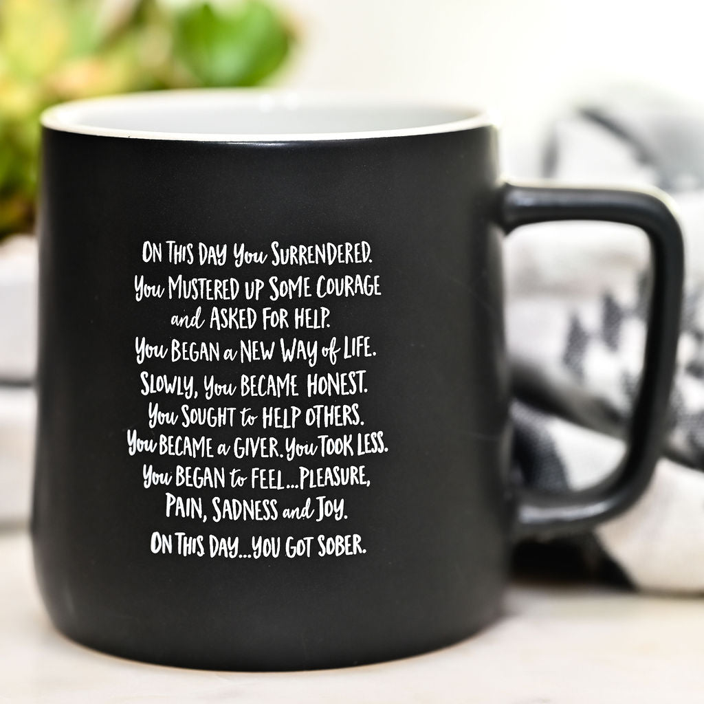 Matte black 11 oz ceramic sobriety mug with a white interior, printed with the full "On This Day You Got Sober" poem in white handwritten lettering, styled on a white counter with a succulent and a black and white checked cloth.