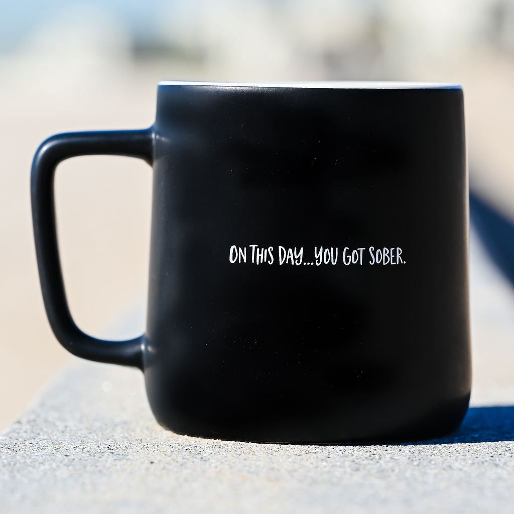 Reverse side of the matte black sobriety mug, printed with the single line "On this day… you got sober." in white handwritten lettering, resting on grey stone in daylight.