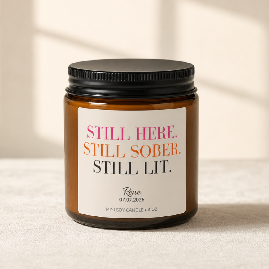 An amber glass jar candle with a black lid and a cream label reading 'STILL HERE. STILL SOBER.