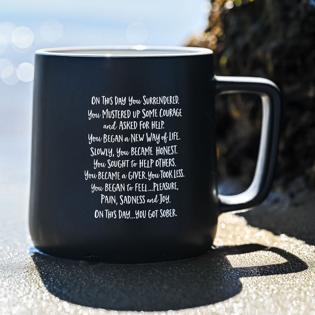 Matte black sobriety mug printed with the full "On This Day You Got Sober" poem, sitting on sunlit sand near the water with a long morning shadow.