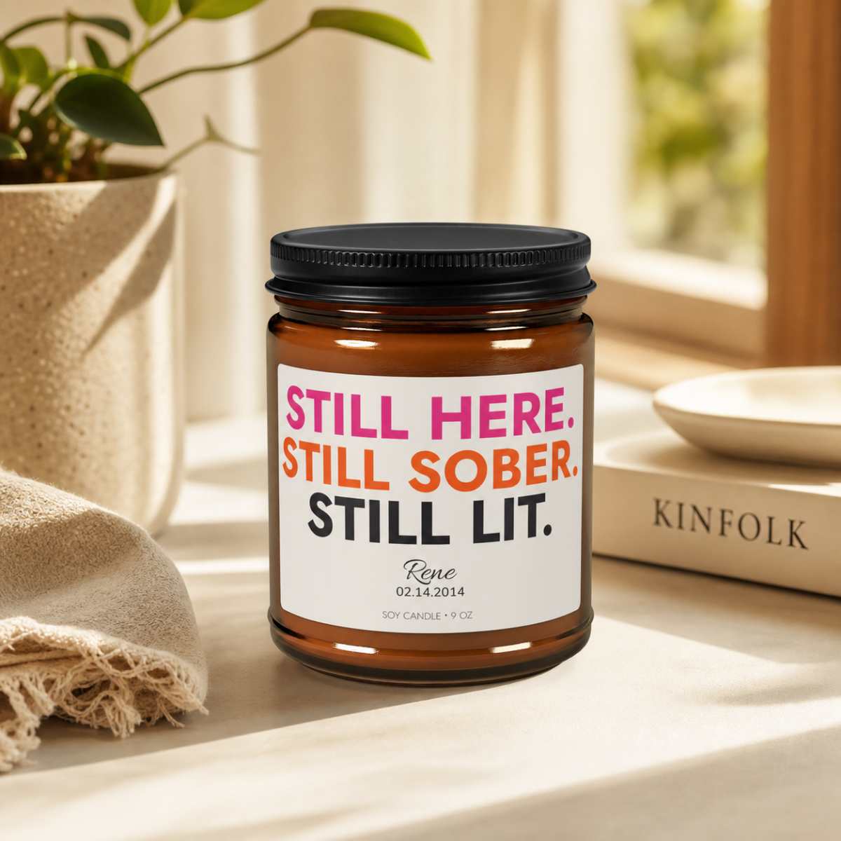 Personalized sobriety candle in a 9 oz jar styled on a windowsill with a plant and a Kinfolk book
