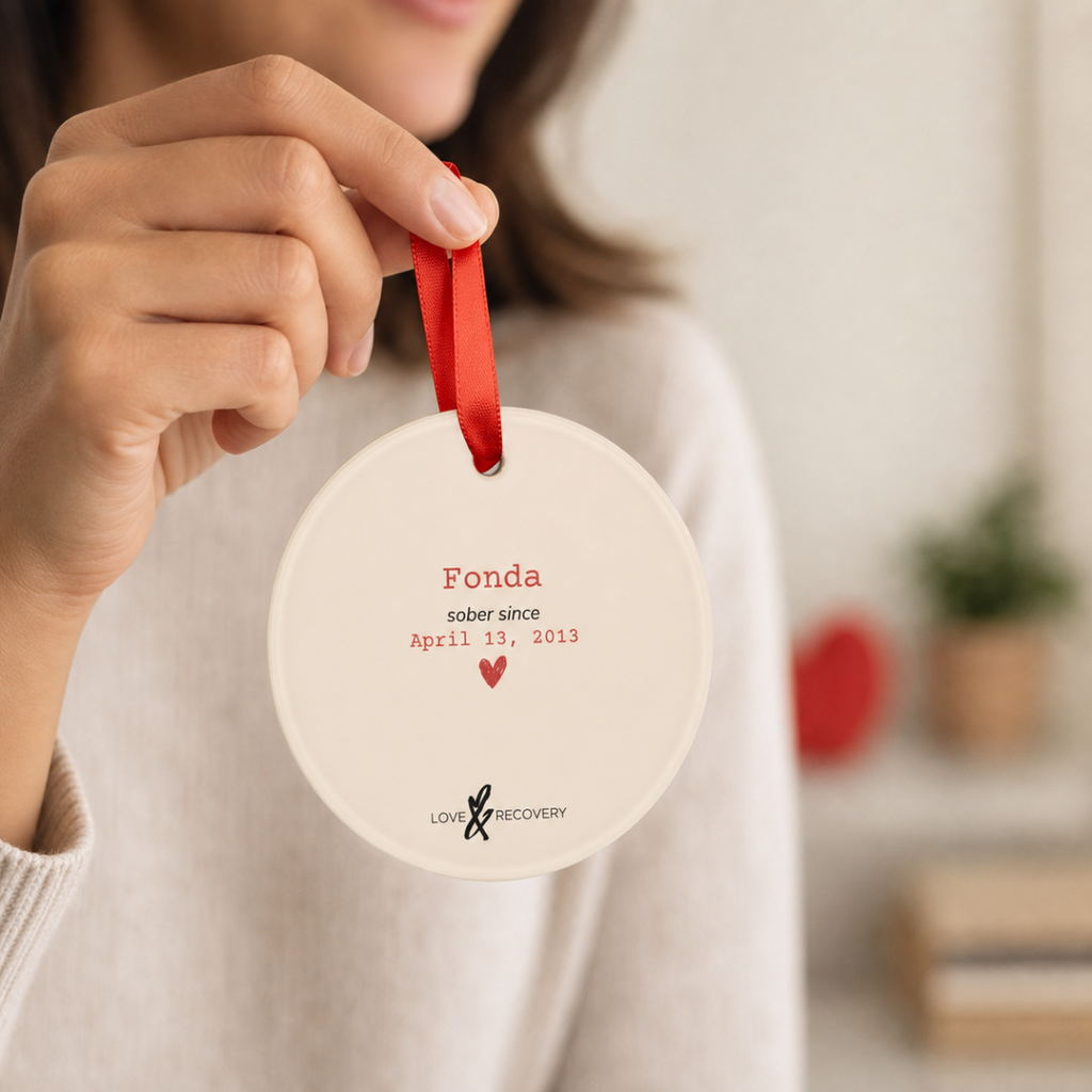 Hand holding the personalized sobriety ornament by its red ribbon, showing the custom name and sober date