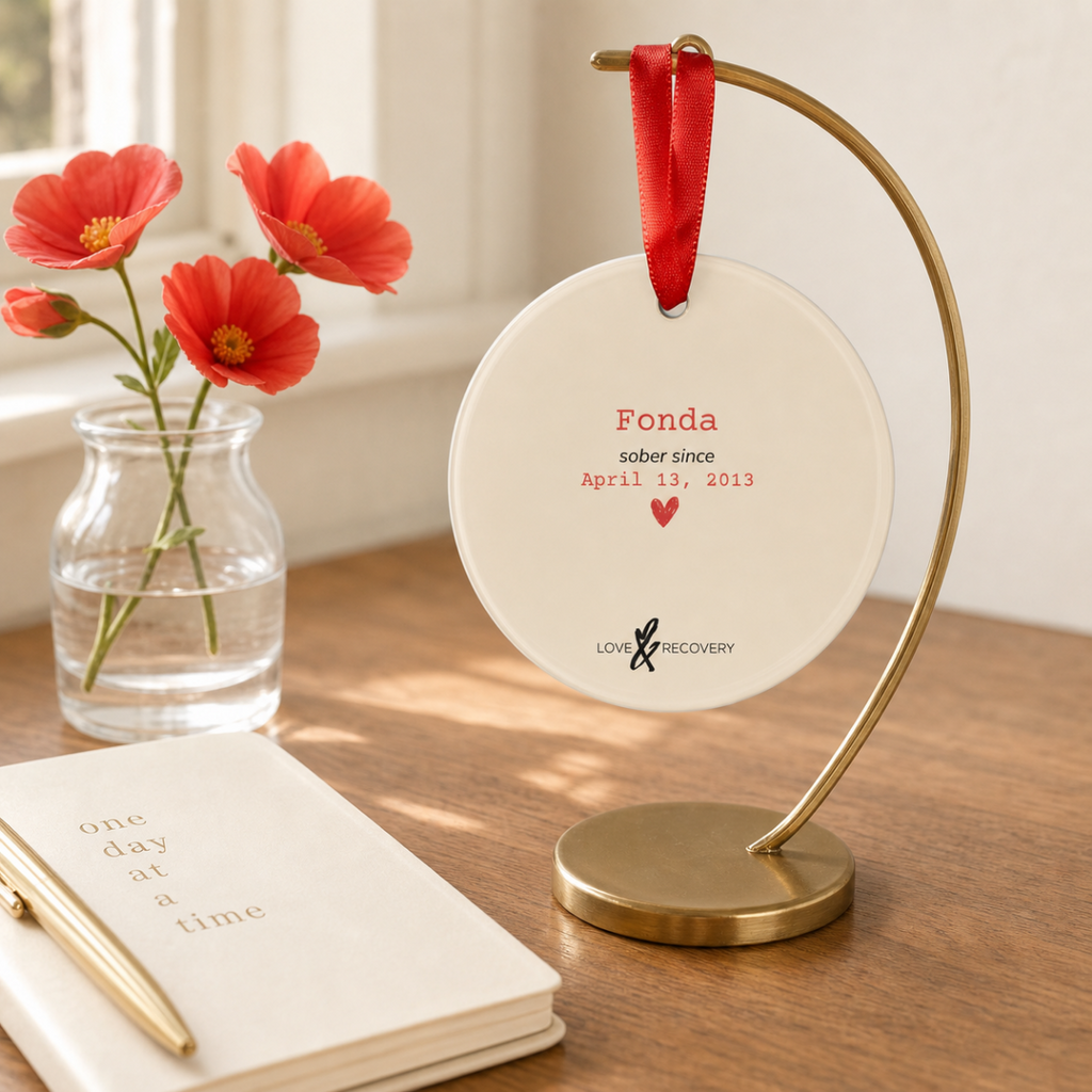 Personalized sobriety ornament displayed year-round on a brass stand beside red flowers and a journal.