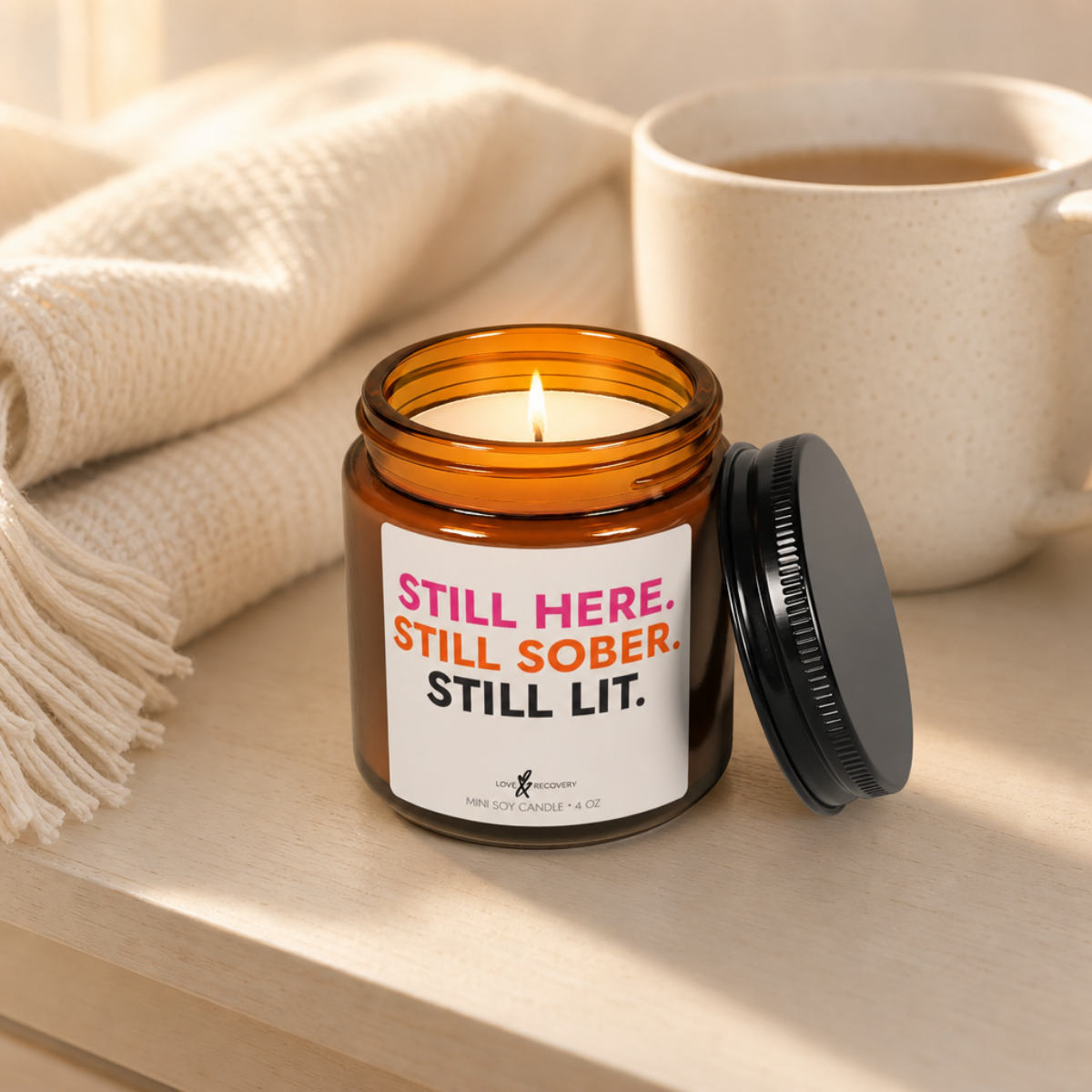 Lit sobriety candle in a 4 oz jar styled with a cream blanket and a mug of coffee
