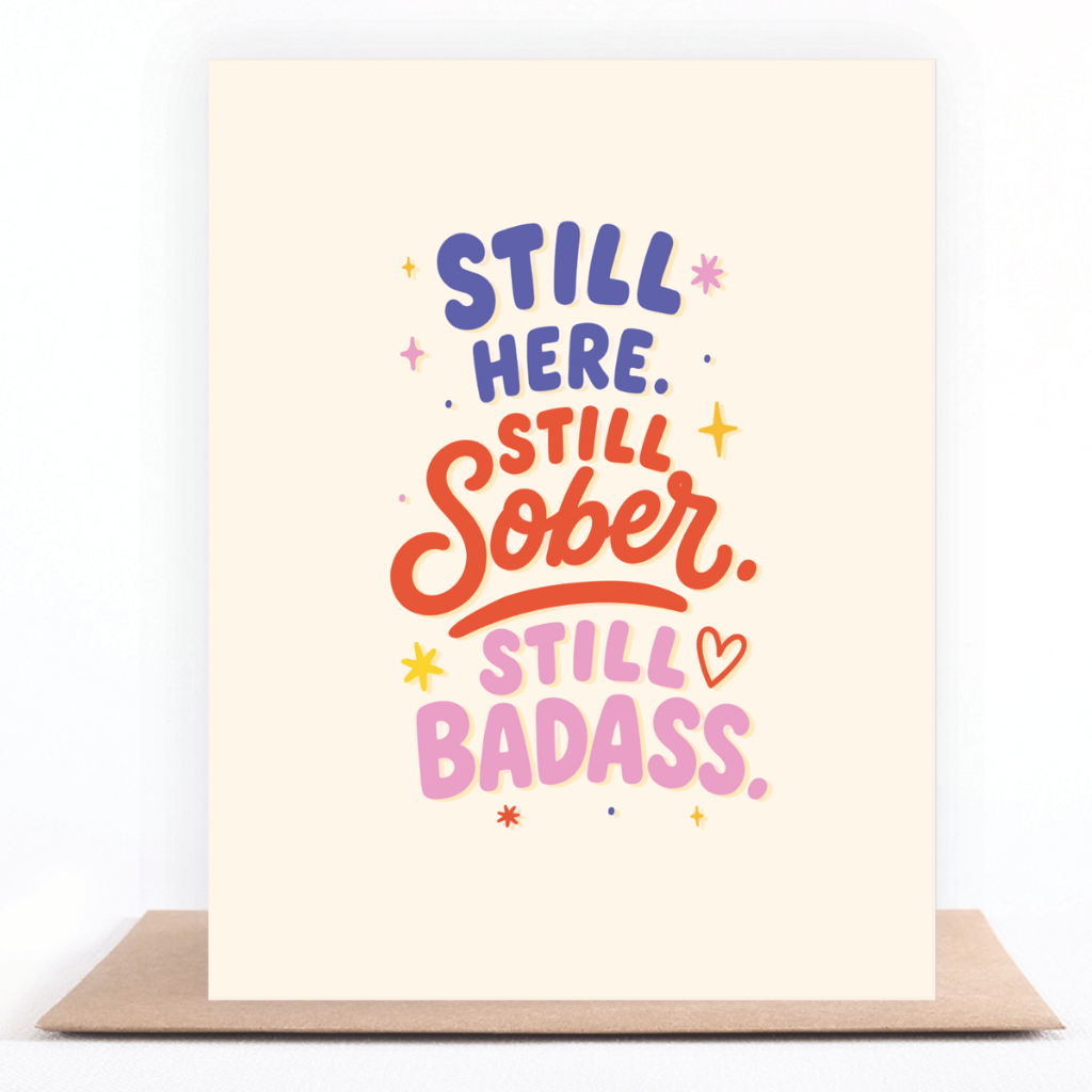 Colorful sobriety card reading “Still Here. Still Sober. Still Badass.” displayed with a kraft envelope.