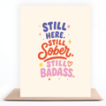 Colorful sobriety card reading “Still Here. Still Sober. Still Badass.” displayed with a kraft envelope.